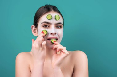 Çamur maskeli ve yüzünde salatalıklar olan çıplak genç bir kadın, maviden soyutlanmış. Güzel bir model kozmetik krem sürüyor. Yüz bakımı. Çıplak omuzlu üstsüz kadın yüz kremi sürüyor.