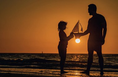 Baba oğul oyuncak yelkenli tekneyle, oyuncak gemiyle oynuyorlar. Seyahat ve macera konsepti. Baba ve oğul yaz günbatımı kumsalında. Baba ve oğul gün batımında plajda oynuyorlar. Erkek aile. Babalar Günü