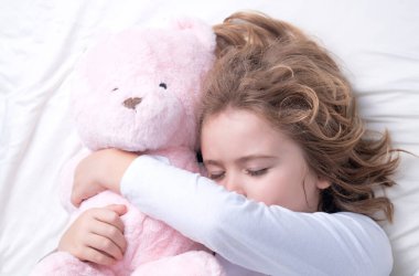 Günlük uyku. Oyuncak ayıyla yatakta uyuyan bir çocuk. Battaniyenin altında uyuyan tatlı çocuk. Çocuk yastığa uzanmış. Çocuk uyuyor, sağlıklı bir uyku çekiyor ya da kestiriyor.