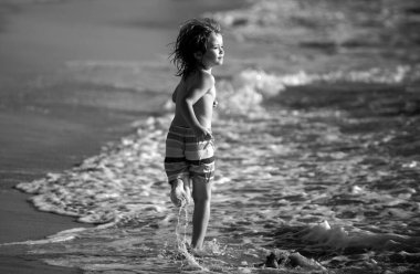 Kumsalda koşan küçük çocuk mavi denizde su sıçratıyor. Çocuk yaz plajında yürüyor.