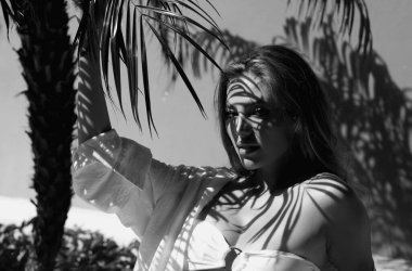 Yüzünde palmiye gölgesi olan seksi bir kadın. Bronzlaşmış kadın dinleniyor ve güneşleniyor. Seksi mayo bikinili, baştan çıkarıcı sarışın kadın. Mükemmel bronzlaşmış ince vücut. Yaz tropik havası