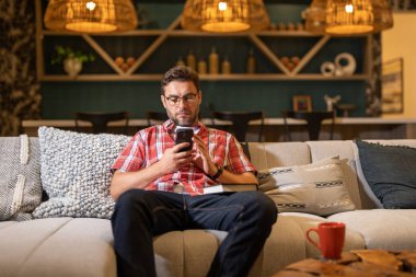 Evdeki kanepede cep telefonu kullanan yakışıklı, neşeli adam. Çekici beyaz bir adam gece yarısı evdeki kanepede dinleniyor telefonla mesaj atıyor, akıllı telefondan internette sörf yapıyor.