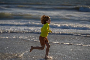 Gün doğumunda sahilde koşan küçük bir çocuk. Deniz sahilinde koşan mutlu çocuk. Yaz günü okyanus kenarında koşan sevimli bir çocuk. Çocuklar tropikal plajda koşuyor. Çocuk su sıçratarak oynuyor.
