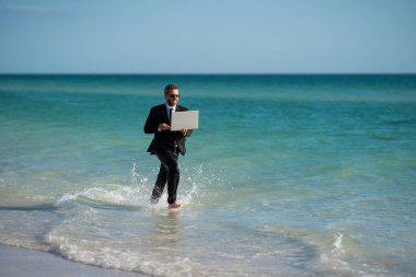 Kumsalda yalın ayak koşan bir iş adamı kıyafeti. Yaz tatilindeki yakışıklı iş adamı. Elinde dizüstü bilgisayarla denizde koşan bir iş adamı. İş adamı deniz suyuna atladığı için heyecanlı. Çılgın işadamı