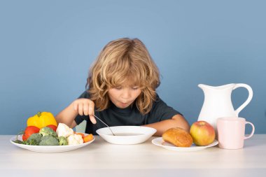 Küçük şirin bir çocuk masada yemek yiyor, kasede çorba tadıyor. Mutlu gülümseyen çocuk sağlıklı ev yemekleri yiyor.