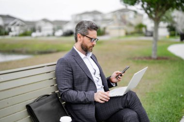 Parktaki bankta oturan bir iş adamı. Adam dışarıda bilgisayarla uğraşıyor. Parktaki bankta oturup dizüstü bilgisayarla online iş yapan yakışıklı bir iş adamı. Takım elbiseli bir iş adamı parkta haber okuyor.