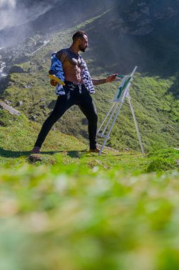 Açık havada çizim yapan İspanyol sanatçı kaslı adam. Resim sanatı. Erkek sanatçı Alp dağlarında resim çizer. Fırçayla ya da akrilik boyayla resim yapan adam. Erkek sanatçı tuvale bir resim çizer.