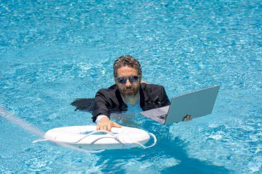 Yaz işlerine yardım ediyorum. Takım elbiseli iş adamı dizüstü bilgisayarı ve deniz kıyısındaki suda can simidi tutuyor. Kurtarma işadamı. İş adamına yardım etmek. İşi kurtar. Yaz krizi. Çılgın yaz. Sıcak iş yazı