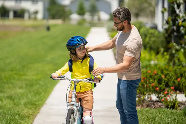 Baba oğul parkta bisiklete biniyor. Babası yaz günü bisiklet sürerken kasklı çocuk. Baba oğluna bisiklete binmeyi öğretiyor. Bisiklet kasklı çocuk ve bisikletli baba. Babalar Günü