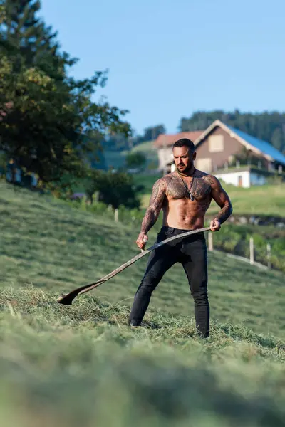 stock image Strong farmer using scythe. Sexy Farmer with a scythe on green grass field. Muscular Farmer cut grass. Rural farmer on summer meadow mowing grass with classic traditional scythe