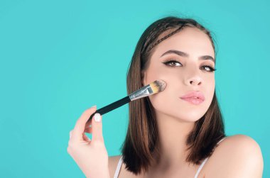 Beautiful young woman applying makeup on studio background. Pretty girl holding makeup brushes and make up on face with cosmetics. Facial beauty. Perfect skin, natural make up