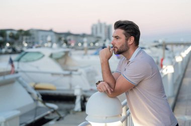 Rich businessman dreaming and thinking near the yacht. Portrait of stylish male model outdoor. Fashion male posing near skyscraper on the street. Fashionable man in urban style