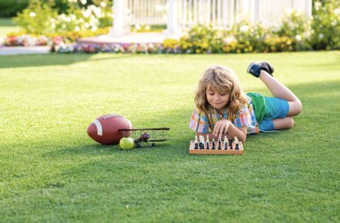 Çocuk satranç oynuyor ve arka bahçede ya da yaz parkında eğleniyor. Açık havada satranç okulu. Çocuk, yaz parkında çimlerin üzerinde yatarak satranç oynamayı düşünüyor ya da planlıyor. Zeki ve zeki okul çocukları.