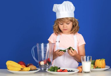 Şef şapkalı ve önlüklü çocuk yemek hazırlıyor. Mutfakta sebzeli küçük bir aşçı. Doğal çocuk yemekleri.