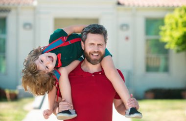 Baba, okul sonrası oğlunu sırtında taşıyor. Sırt çantalı ilkokul öğrencisinin ebeveyni ve öğrencisi.