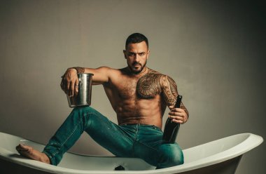 Banyoda seksi vücudu olan kot pantolonlu, sakalsız yakışıklı bir adam. Banyodaki seksi maço adam. Şampanya şişesi tutan ve banyoda poz veren güçlü kaslı dövmeli adam.