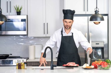 Mutfakta yemek pişiren yakışıklı bir adam. Mutfakta sebze ve etle yemek pişiren olgun bir adam. Kafkasyalı önlüklü adam ve aşçı şapkalı aşçı mutfakta lezzetli bir öğle yemeği pişiriyor. Akşam yemeği için sağlıklı yemek.