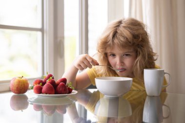 Tatlı komik çocuk kasede az yağlı soya sütü yiyor. Sağlıklı beslenmek için. Evde kahvaltı yapan sevimli küçük bir çocuk. Kaşık ve kase taşıyan çocuk yemek masasında yemek yiyor.