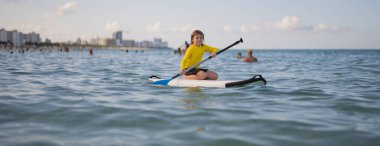 Çocuk ayakta raket üzerinde yüzüyor. Su sporları, aktif yaşam tarzı. Okyanusta kürek çeken bir çocuk var. Çocuk Paddle binicisi. Yaz Suyu sporu, SUP sörfü. Yaz tatili