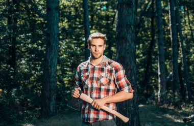 A handsome young man with a beard carries a tree. Stylish young man posing like lumberjack. Lumberjack worker walking in the forest with axe clipart