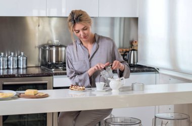 Mutfakta kahveye süt koyan bir kadın. Sabahları seksi ev hanımı