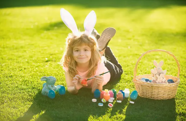 stock image Child boy with easter eggs and bunny ears laying on grass painting eggs