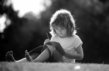 Komik çocuk ödevini yapıyor ve parkta çim üzerinde kitap okuyor. Çocuklar için uzaktan eğitim. Anaokulu öğrencisi