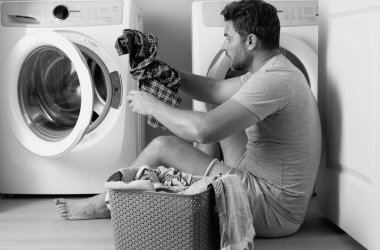 Çamaşır makinesinin yanında elbiseli bir adam. Yakışıklı adam çamaşır makinesinin önünde oturur. Çamaşır makinesini kirli çamaşırlarla dolduruyor. Adam elbiseleri temizliyor. Yalnız bir adam için ev işi. Ev çamaşırları