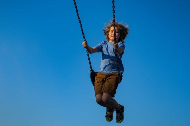 Zincir sallanan çocuk, mavi gökyüzüne karşı eğleniyor. Tasasız ve özgür çocukluk. Açık havada oynayan aktif bir çocuk. Çocuk parkında. Yazın dışarıda oyun parkında eğlenen sevimli bir çocuk. Çocuk sallanıyor