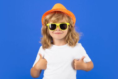 Stüdyoda izole edilmiş arka planda onay veren bir çocuk. Mavi stüdyoda, şapkalı, tişörtlü ve güneş gözlüklü şirin bir çocuğun moda portresi. Çocuk işaret veriyor.