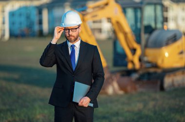 İnşaat işçisi yatırımcı. İnşaat alanının önündeki yatırımcı. Modern ev inşaatında başarılı yakışıklı bir adam. Takım elbiseli ve kasklı orta yaşlı yatırımcının portresi