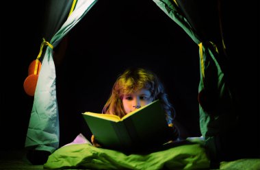 Çocuklar çadırda kitap okuyor. Kitapla çocuk okuma hikayesi. Çocukların yüzü gece lambasıyla kaplıdır. Eğitim ve okuma kavramı. Çocukların hayal gücünün gelişimi.