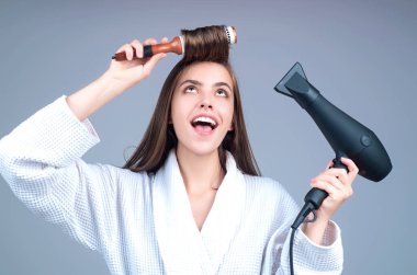 Woman in bathrobe combing hair, drying hair with hairdryer. Portrait of female model with a comb brushing hair. Girl with hairs brush and blow dryer. Hair care and beauty. Morning routine