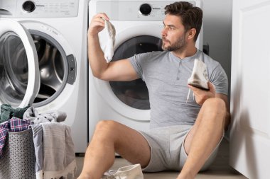 Çamaşır makinesinin yanında elbiseli bir adam. Yakışıklı adam çamaşır makinesinin önünde oturur. Çamaşırhanede çamaşır makinası var. Adam elbiseleri temizliyor. Yalnız bir adam için ev işi. Ev çamaşırları