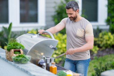 Yakışıklı, gülümseyen İspanyol adam arka bahçede barbekü yapıyor. Piknik barbekü partisi. Barbekü ızgarası için çatal bıçaklı aşçı şefi. Tatil pikniğinde barbekü. Adam barbeküde biftek pişiriyor.