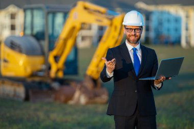 İnşaat işçisi yatırımcı. İnşaat alanının önündeki yatırımcı. Modern ev inşaatında başarılı yakışıklı bir adam. Takım elbiseli ve kasklı orta yaşlı yatırımcının portresi