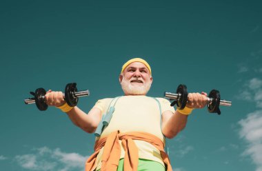 Spor salonundaki son sınıf öğrencisi ağırlık çalışıyor. Kıdemli adam dambılı kaldırıyor. Harekete geçin. Yaz ve aktif tatiller. Büyükbaba sporcu portresi mavi arka planda