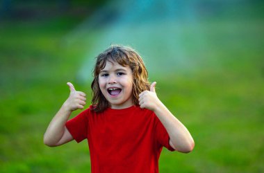 Yaz günü bahçede çimenleri sulayan sevimli küçük bir çocuk. Çocuk yaz bahçesinde oynuyor. Şirin küçük çocuk gülüyor ve su püskürtme fıskiyelerinin altında koşarken eğleniyor. Çimenleri sulamak