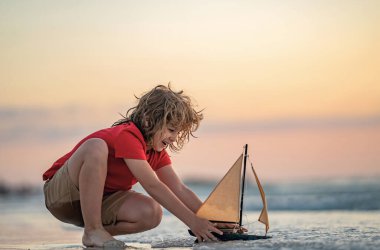 Küçük çocuk yaz tatilinde sahildeki deniz dalgalarında oyuncak tekneyle oynuyor. Çocukluk ve yaz tatili. Seyahatte rüya gör. Yelkenli gemi turu. Çocuklar rüya görür