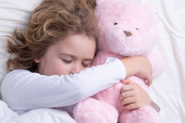 Çocuk gün içinde oyuncak ayıyla yatakta uyuyor. Çocuk yatakta uyuyor. Çocuk battaniyenin altında uyuyor, yastığın üzerinde yatıyor. Çocuk uyuyor, sağlıklı bir uyku çekiyor ya da kestiriyor.