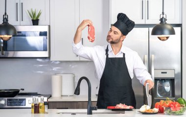Mutfakta yakışıklı bir adam sağlıklı meyve salatası ve et hazırlıyor. Önlüklü bir adam ve aşçı şapkası mutfakta yemek hazırlıyor. Aşçı şapkası aşçısı mutfakta lezzetli yemekler pişiriyor. Modern mutfak iç mimarisi