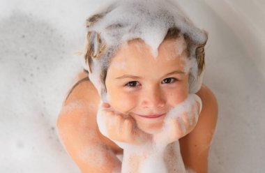 Çocuk şampuanı. Şampuan köpüğü ve banyo yapan saçlarında kabarcıklar olan bir çocuk. Köpüklü çocuk suratı, saç bakımı ve hijyen konsepti. Çocuklar uzun saçlar için şampuan kullanırlar. Banyoda saç yıkamak.