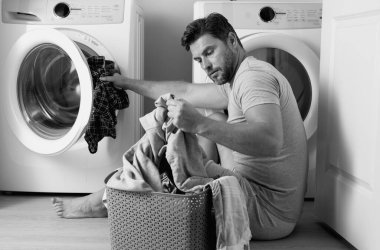 Çamaşır makinesinin yanında elbiseli bir adam. Yakışıklı adam çamaşır makinesinin önünde oturur. Çamaşır makinesini kirli çamaşırlarla dolduruyor. Adam elbiseleri temizliyor. Yalnız bir adam için ev işi. Ev çamaşırları
