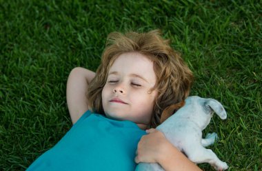 Bahçenin çimlerinde yatan küçük çocuk yavru köpeğe sarılıyor.