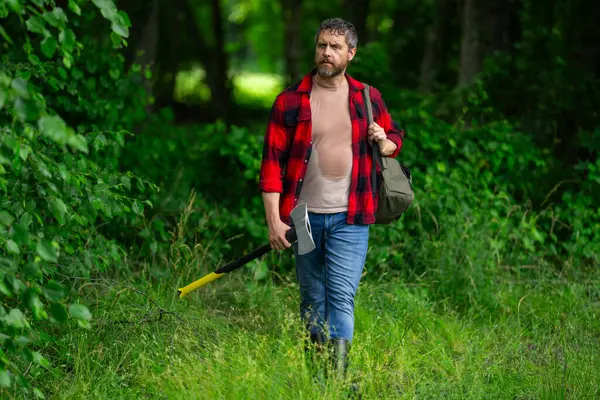 Stock image Mature Lumberjack with serious face holds axe in the forest. A lumberjack with an ax examines the tree before felling. Serious lumberjack. Millennial middle aged man holding a big axe outdoor