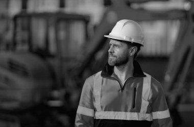 İnşaat alanında miğferli bir işçi. İnsan kazıcı buldozer işçisi. Arkasında kazıcı olan inşaat işçisi. İnşaat işçisi traktörlü ya da inşaatta çalışıyor.