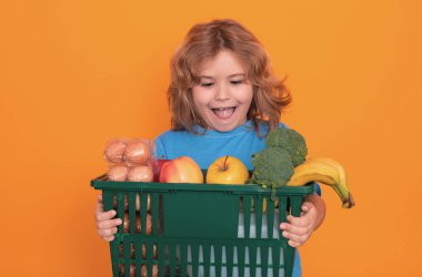 Çocuk bakkal arabası, stüdyo sarı arka plan ve fotokopi alanı. Küçük alışverişçi. Çocuk alışveriş sepetini tutuyor. Markette ya da süpermarkette taze organik, sebze ve meyveler