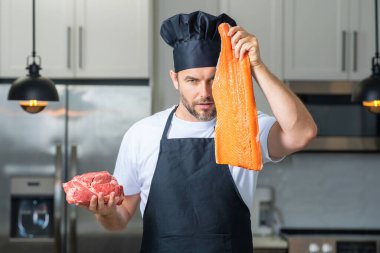 Üniformalı şef mutfakta çiğ et ve balık fileto pişiriyor. Restoran menüsü. İş üniformalı, siyah önlüklü, şef şapkalı bir aşçı. Balık ve et veya somon ve sığır eti doğal proteindir.