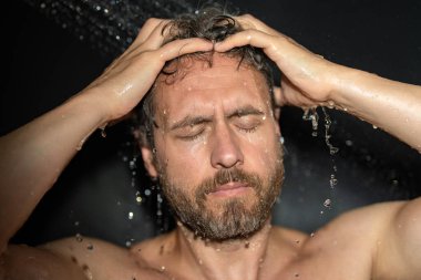 Adam duş alıyor. Adam duş başlığından düşen saçlarını şampuanla yıkıyor. Sabah rutini duş alan adam. Vücut bakımı hijyen duşu. Adam duşta su damlası altında. Sağlık ve kaplıca