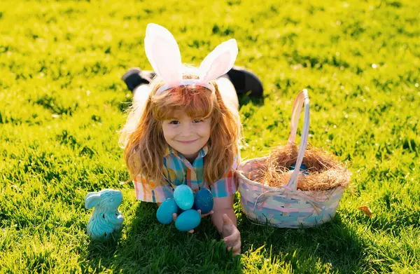stock image Kids boy hunting easter eggs. Child in bunny ears gathering Easter eggs, laying on grass. Spring holiday